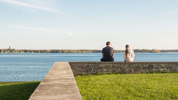 A young couple is sitting on a stone wall overlooking a tranquil lake. They are gazing out at the stunning view and enjoying the peace and quiet, surrounded by nature s beauty and serenity.