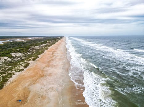 The beach is wide and sandy with a gentle slope into the ocean. The water is clear and blue-green. The sky is blue with scattered clouds. There are dunes covered with sea oats behind the beach.