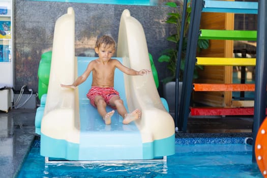 Wet boy in red swim shorts sliding down a plastic water slide into a swimming pool with colorful stairs in the background