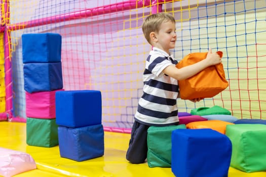 Young boy in striped shirt stacking colorful soft foam cubes inside an indoor play area with bright safety nets and padded floor
