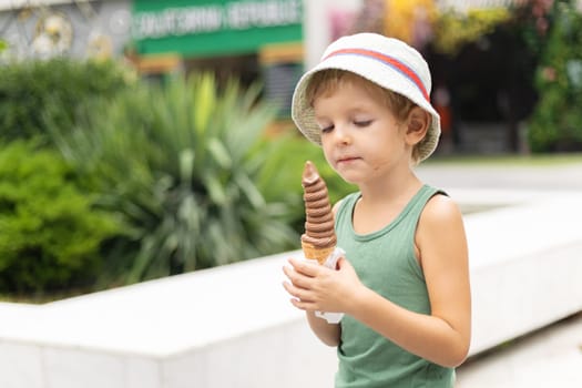 Young child in green shirt and sunhat holding and eating a chocolate soft-serve ice cream cone while standing near a garden walkway