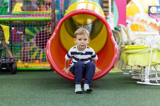 Boy in striped shirt emerging from red and yellow tube slide onto artificial green grass in indoor playground. Studio lifestyle photography. Children activity and amusement concept