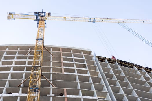 Concrete frame of a multi-story building under construction with large yellow and blue cranes operating against a clear sky background
