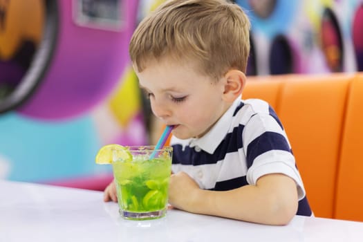 Boy in striped shirt drinking green mocktail with lemon slice through straw while sitting at white table in colorful indoor setting. Studio lifestyle photography. Children and leisure concept