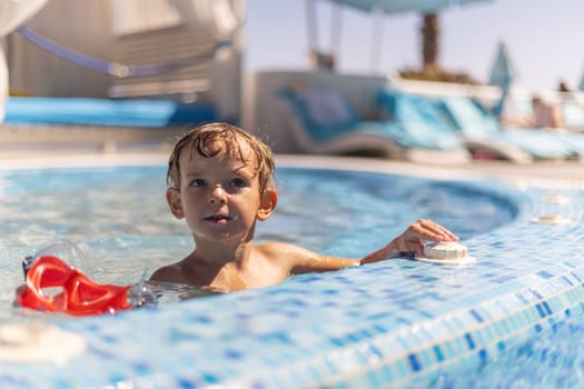 Young boy in a tiled swimming pool resting one arm on the edge and looking to the side, red snorkel mask floating nearby, blurred sun loungers in background