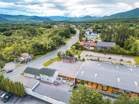 This stunning aerial view captures a charming New England town nestled among majestic mountains. Lush green forests surround the area, with a clear blue sky and fluffy clouds enhancing the beauty.