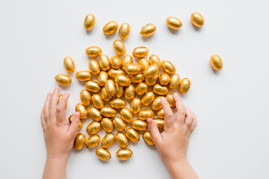 Child hands with golden wrapped chocolate eggs on white background.