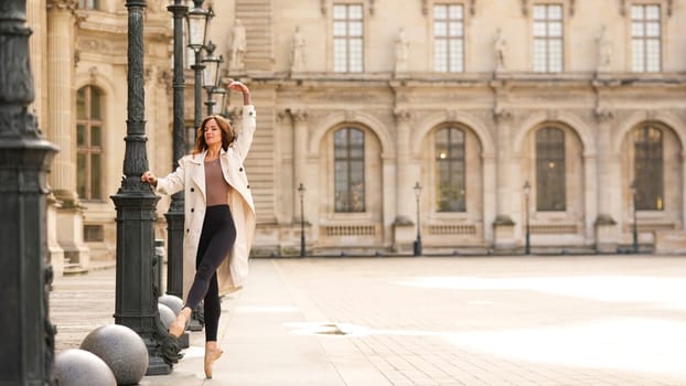 PARIS, FRANCE 11 APRIL 2025 A ballet dancer in a beige trench coat and pointe shoes performs a graceful movement near the historic Louvre Museum