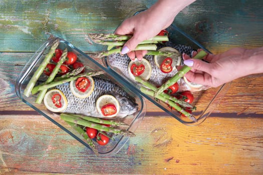 womans hand puts green asparagus on fresh large dorado fish, tilapia, carp in a baking dish with cherry tomatoes,step by step recipe instructions, top view on an old wooden table, step six