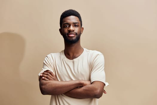 A confident young African man wearing a beige t shirt stands with his arms crossed against a warm background, exuding self assurance and calmness