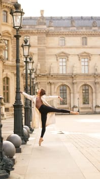 A ballet dancer wearing a trench coat and pointe shoes performs a graceful pose near the historic Louvre Museum in Paris. The building is one of the most visited landmarks