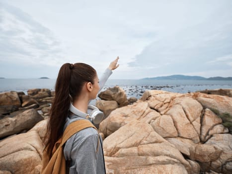 A young woman in casual outdoor clothing stands on rocks at a beach, pointing towards the distant horizon She looks curious and contemplative, enjoying the tranquility by the ocean