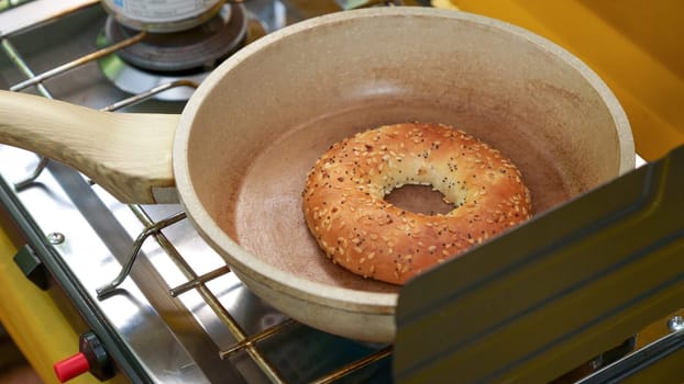 The bread is a bagel with sesame seeds on top. The ceramic pan is brown and has a wooden handle. The camp stove is silver and has a black grate on top. The bagel is round and has a hole in the middle.
