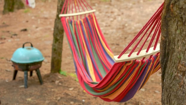 A vibrant, colorful striped hammock is gracefully hanging between two tall trees in a peaceful forest. The hammock remains empty, gently swaying back and forth in the soft breeze, inviting relaxation.