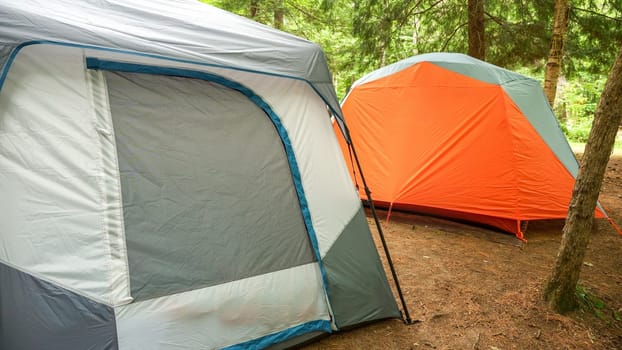 Two colorful camping tents are set up in the lush green woods, surrounded by tall trees. The tents are pitched on a level patch of ground, providing a cozy outdoor space for camping enthusiasts.