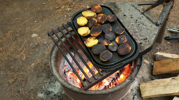 A close-up of rustic potatoes cooking on a campfire grill grate, highlighting their charred and crispy exterior while revealing the soft and fluffy interior, perfect for outdoor cooking enthusiasts.