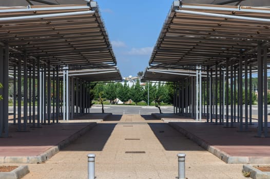 Canopy structures create a geometric pattern in an open area, bathed in bright sunlight. Sparse trees line the backdrop, adding a touch of nature to the urban space.