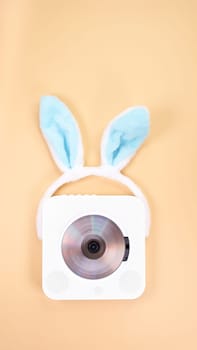 The image features a vintage white portable CD player, elegantly designed, adorned with a playful pair of blue bunny ears headband. Set against a soft peach color background, it evokes nostalgia.