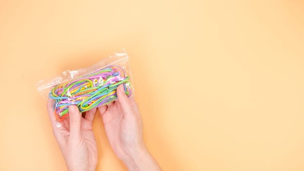A hand holding a transparent plastic bag filled with colorful rubber bands against a peach background. The rubber bands are in various colors, including pink, blue, green, and yellow.