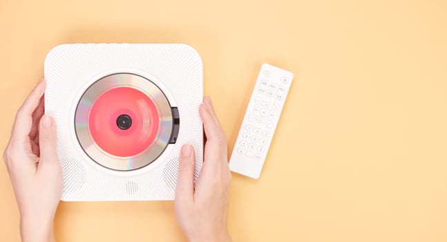 The image shows a white portable CD player featuring a remote control resting on a peach background. Inside the CD player is a vibrant red CD, showcasing its modern design and functionality.