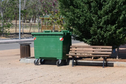 A vibrant green dumpster sits beside a weathered wooden bench in a peaceful park. Sunlight filters through the leaves as distant blooms add color to the scene. Quietness envelops this inviting spot.