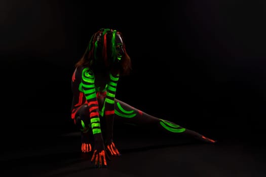 Full body of anonymous female with bright neon red and green fluorescent paints on naked body sitting on haunches in dark studio