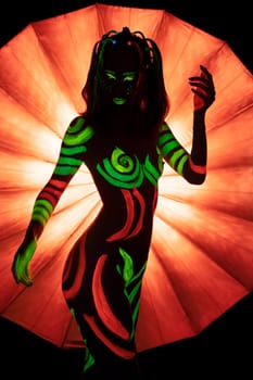 Unrecognizable naked female with bright green and red fluorescent paints on body standing against bright orange umbrella