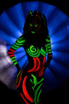 Unrecognizable naked female with bright green and red fluorescent paints on body standing against blue background in darkness