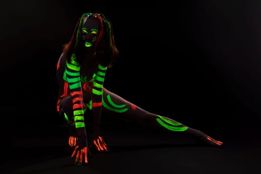 Full body of anonymous female with bright neon red and green fluorescent paints on naked body sitting on haunches in dark studio