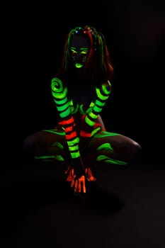 Full body of anonymous female with bright neon red and green fluorescent paints on naked body sitting on haunches in dark studio