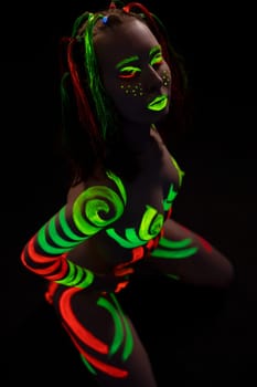 anonymous topless woman with bright neon red and green fluorescent bodyart on body and face stand on knees in dark