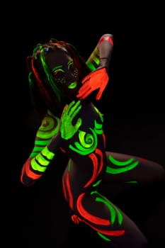 From above of unrecognizable nude lady with creative face and body art made with bright fluorescent paints sitting on haunches in darkness