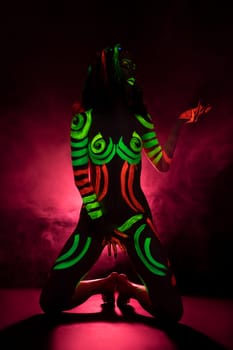 Full length of topless female model with bright UV paints on body and face sitting on haunches against pink smoke background
