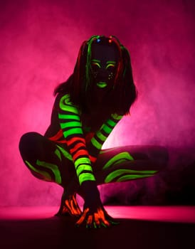 Full length of unrecognizable female model with long hair and bright UV paints on body and face sitting on haunches against pink background