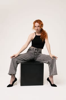 self assured young female model with curly red hair in trendy outfit sitting on chair with legs apart and looking at camera