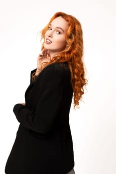 Side view of self assured young female model with long curly red hair in classy outfit smiling and looking at camera against white background