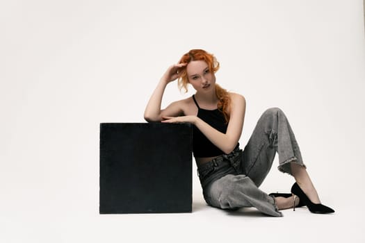 Full body of self assured young female model with curly red hair in trendy outfit sitting on ground with legs apart