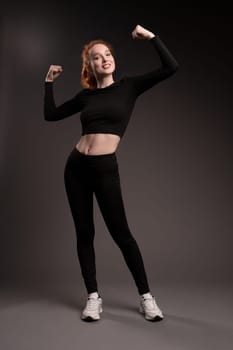 Full body of confident young fit female athlete with long curly red hair in black sportswear show arms and smile on camera