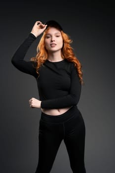 Self assured young female model with curly long red hair in trendy activewear adjusting cap and looking at camera while leaning forward against gray background