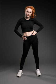 Full body of confident young fit female athlete with long curly red hair in black sportswear and sneakers standing with hands on waist