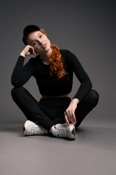 Full body of relaxed young athletic woman with long ginger hair in stylish sportswear and sneakers sitting on floor and looking at camera