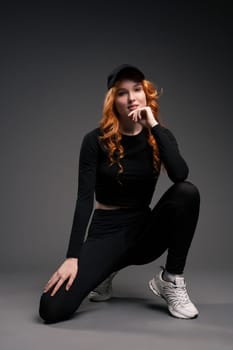 Self assured young female model with curly long red hair in trendy activewear adjusting cap and looking at camera while leaning forward against gray background