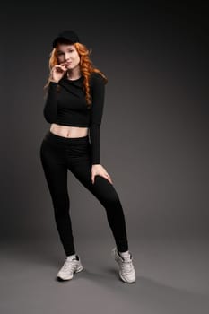 Self assured young female model with curly long red hair in trendy activewear adjusting cap and looking at camera while leaning forward against gray background