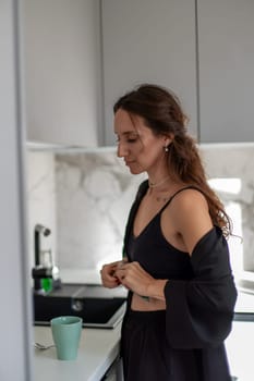 Woman in black lingerie and robe adjusts clothing in a modern kitchen, a teal mug on the counter nearby. Espresso machine visible.