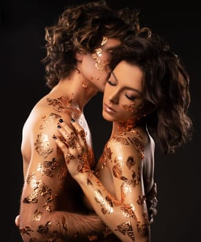 Side view of sensual loving couple in underwear with wavy dark hair and golden foils on bodies embracing gently while standing against black background