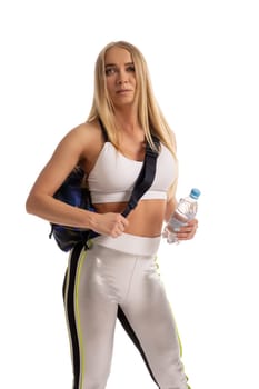 Self assured young female athlete with long blond hair in sportswear and backpack standing against white background with water bottle in hand and looking away