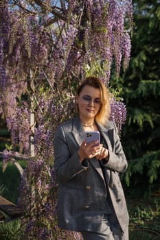 A woman in a grey blazer and skirt stands in front of a blooming wisteria tree, looking at her phone.
