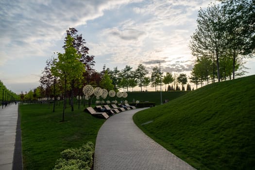 A winding path leads through a grassy park, flanked by trees and a row of lounge chairs. The sky above is a mix of blue and gray clouds.
