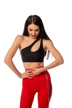Self assured young female athlete with long dark hair in crop top and leggings measuring waist with tape and screaming happily against white background