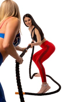 Strong brunette in sportswear with long hair smiling and pulling rope with blond girlfriend during fitness training against white background
